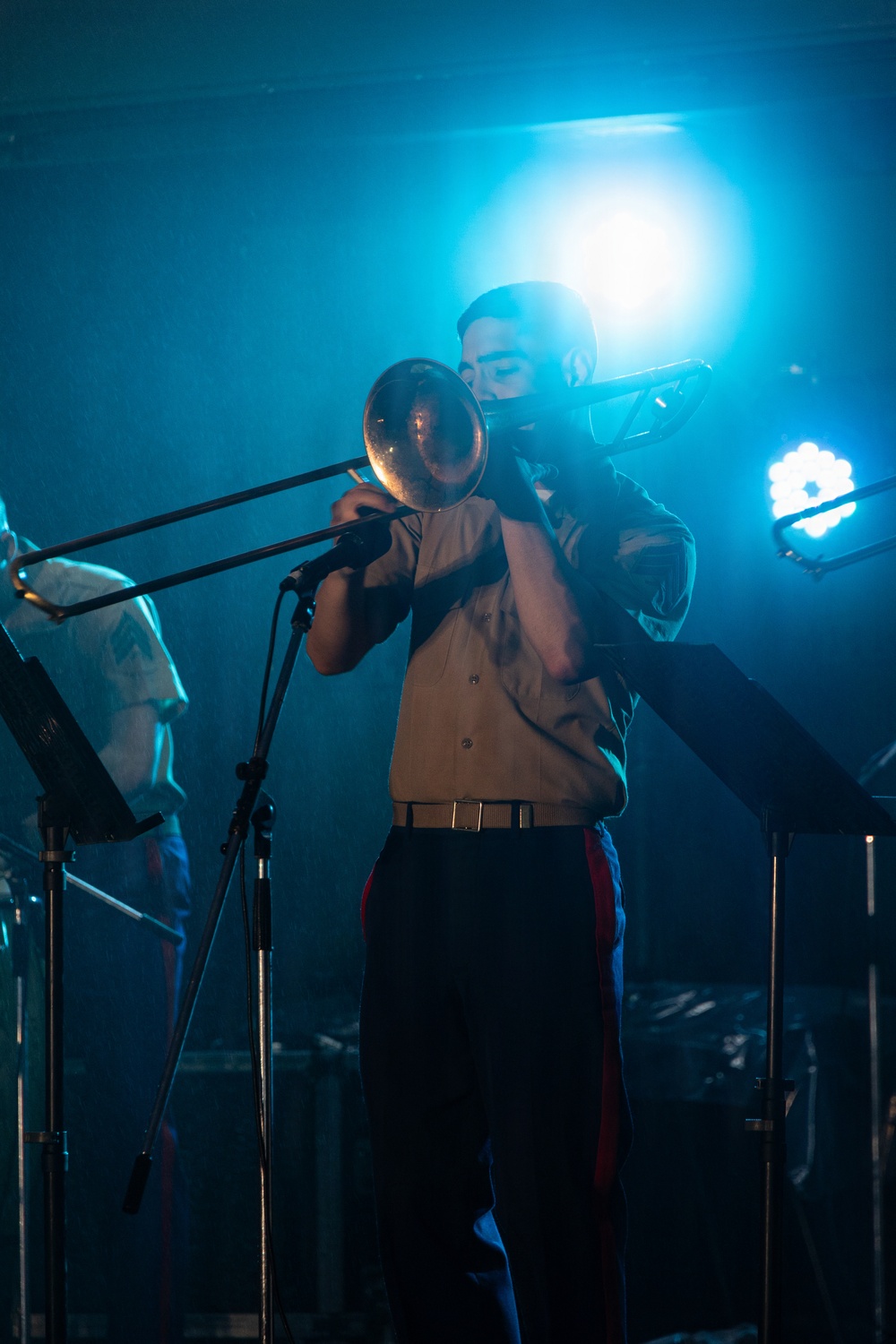 A musician is performing on stage playing a trombone under dramatic blue stage lighting. The performer is dressed in a light-colored button-up shirt with dark trousers featuring a red stripe, and a belt. A music stand and microphone stand are visible in the foreground, with at least one other performer partially visible in the background.