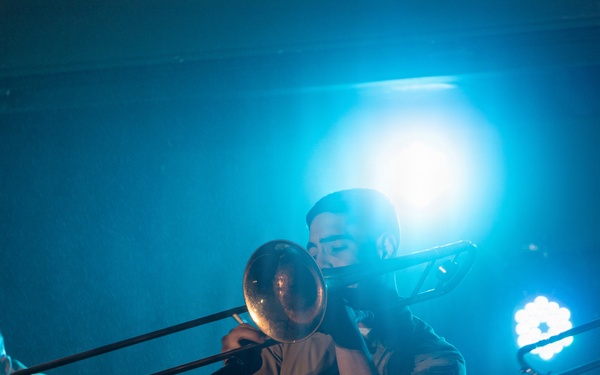 A musician is performing on stage playing a trombone under dramatic blue stage lighting. The performer is dressed in a light-colored button-up shirt with dark trousers featuring a red stripe, and a belt. A music stand and microphone stand are visible in the foreground, with at least one other performer partially visible in the background.