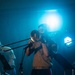 A musician is performing on stage playing a trombone under dramatic blue stage lighting. The performer is dressed in a light-colored button-up shirt with dark trousers featuring a red stripe, and a belt. A music stand and microphone stand are visible in the foreground, with at least one other performer partially visible in the background.