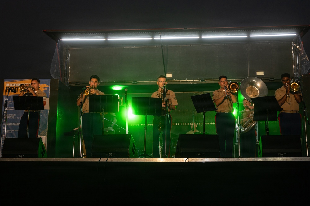 A military band performs on an outdoor stage at night, illuminated by white overhead lights and green stage lighting. The musicians, dressed in tan/khaki military uniforms consistent with U.S. Marine Corps attire, are playing brass and woodwind instruments. A banner on the left references 'Fuji Friendship Festival,' suggesting this is a U.S.-Japan military community event.