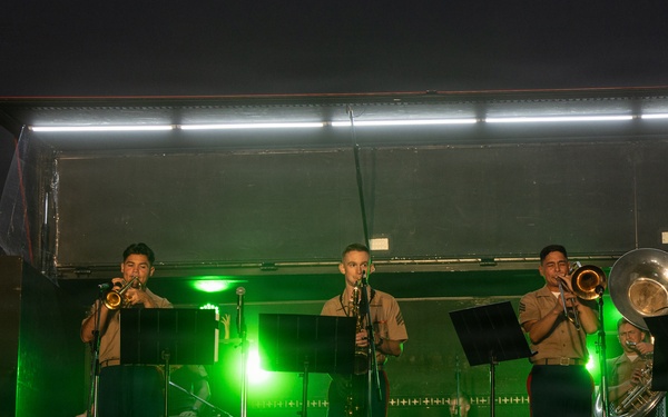 A military band performs on an outdoor stage at night, illuminated by white overhead lights and green stage lighting. The musicians, dressed in tan/khaki military uniforms consistent with U.S. Marine Corps attire, are playing brass and woodwind instruments. A banner on the left references 'Fuji Friendship Festival,' suggesting this is a U.S.-Japan military community event.