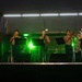 A military band performs on an outdoor stage at night, illuminated by white overhead lights and green stage lighting. The musicians, dressed in tan/khaki military uniforms consistent with U.S. Marine Corps attire, are playing brass and woodwind instruments. A banner on the left references 'Fuji Friendship Festival,' suggesting this is a U.S.-Japan military community event.