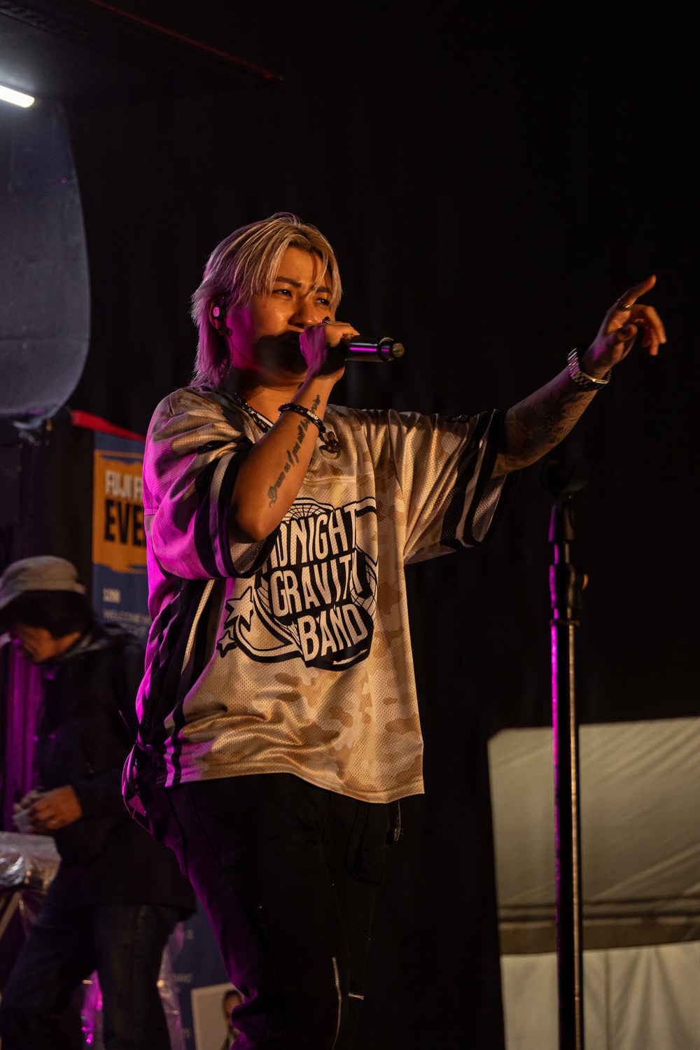 A performer stands on stage holding a microphone and pointing outward toward the audience, wearing a jersey-style shirt reading 'Midnight Gravity Band.' The scene is lit with purple/pink stage lighting against a dark background. The performer has blonde/light-colored hair, visible tattoos on their arms, and wears bracelets.