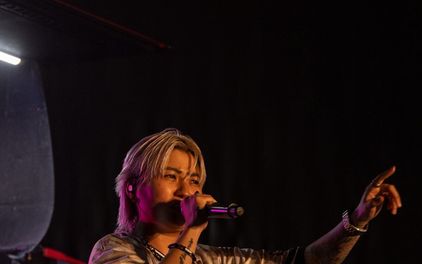 A performer stands on stage holding a microphone and pointing outward toward the audience, wearing a jersey-style shirt reading 'Midnight Gravity Band.' The scene is lit with purple/pink stage lighting against a dark background. The performer has blonde/light-colored hair, visible tattoos on their arms, and wears bracelets.