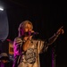 A performer stands on stage holding a microphone and pointing outward toward the audience, wearing a jersey-style shirt reading 'Midnight Gravity Band.' The scene is lit with purple/pink stage lighting against a dark background. The performer has blonde/light-colored hair, visible tattoos on their arms, and wears bracelets.