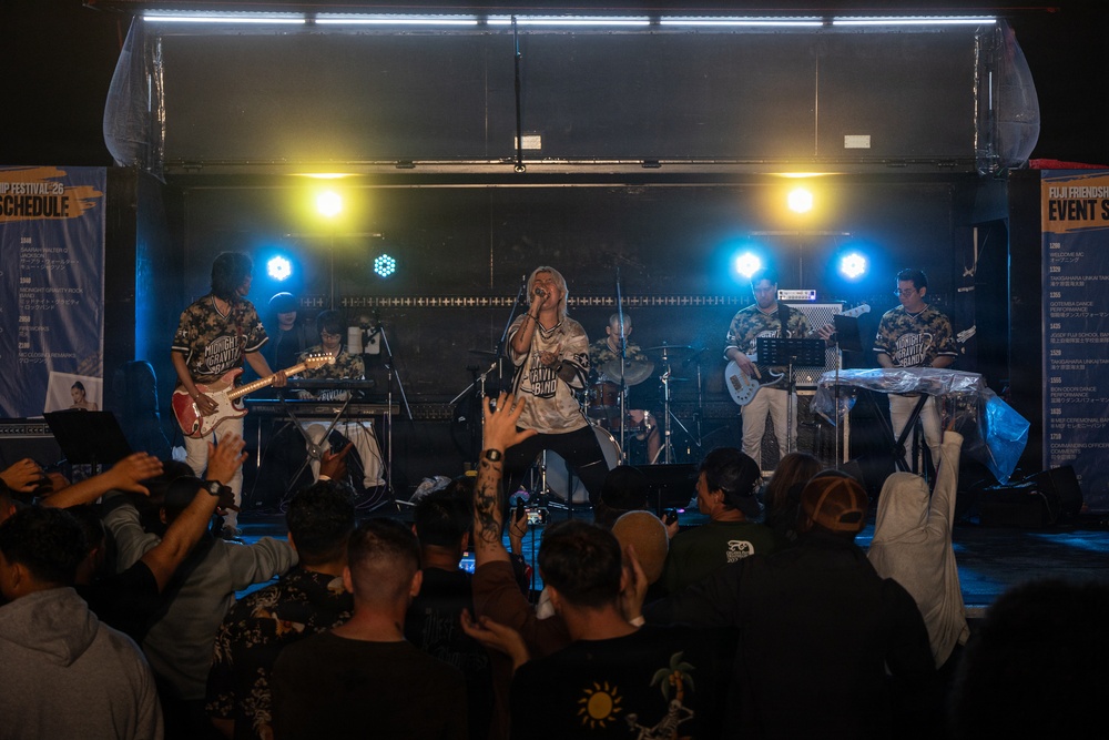 A live band performance is taking place on an indoor stage at what appears to be the 'Fuji Friendship Festival.' The band, identified as 'Midnight Gravity Band' based on visible shirt text and signage, is performing to an engaged crowd. The scene captures an energetic concert atmosphere with stage lighting, multiple musicians, and an enthusiastic audience.