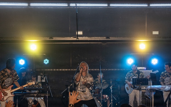A live band performance is taking place on an indoor stage at what appears to be the 'Fuji Friendship Festival.' The band, identified as 'Midnight Gravity Band' based on visible shirt text and signage, is performing to an engaged crowd. The scene captures an energetic concert atmosphere with stage lighting, multiple musicians, and an enthusiastic audience.