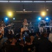 A live band performance is taking place on an indoor stage at what appears to be the 'Fuji Friendship Festival.' The band, identified as 'Midnight Gravity Band' based on visible shirt text and signage, is performing to an engaged crowd. The scene captures an energetic concert atmosphere with stage lighting, multiple musicians, and an enthusiastic audience.