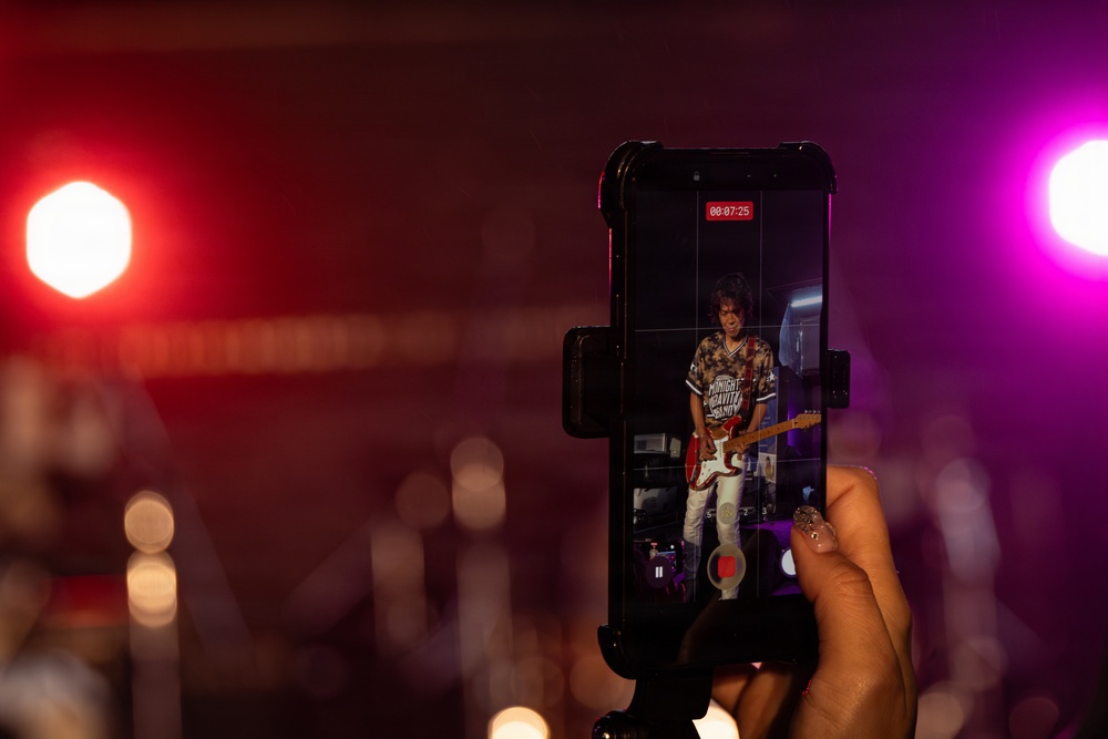 A person is recording a live concert performance on a smartphone mounted in a phone grip/holder. The phone screen displays a guitarist performing on stage, with a recording timer showing 00:07:25. The background shows a dimly lit concert venue with red and purple/magenta stage lighting.