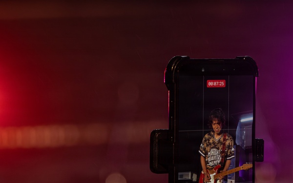 A person is recording a live concert performance on a smartphone mounted in a phone grip/holder. The phone screen displays a guitarist performing on stage, with a recording timer showing 00:07:25. The background shows a dimly lit concert venue with red and purple/magenta stage lighting.