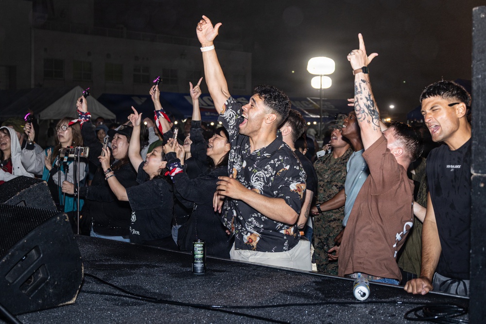 A lively outdoor concert or festival event photographed at night, showing an energetic crowd gathered near the stage front. The audience includes a diverse mix of civilians and at least one person in military camouflage uniform, all visibly engaged and enthusiastic. The scene appears to be at a military base community event or public festival.