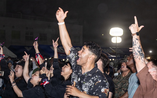 A lively outdoor concert or festival event photographed at night, showing an energetic crowd gathered near the stage front. The audience includes a diverse mix of civilians and at least one person in military camouflage uniform, all visibly engaged and enthusiastic. The scene appears to be at a military base community event or public festival.