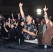 A lively outdoor concert or festival event photographed at night, showing an energetic crowd gathered near the stage front. The audience includes a diverse mix of civilians and at least one person in military camouflage uniform, all visibly engaged and enthusiastic. The scene appears to be at a military base community event or public festival.