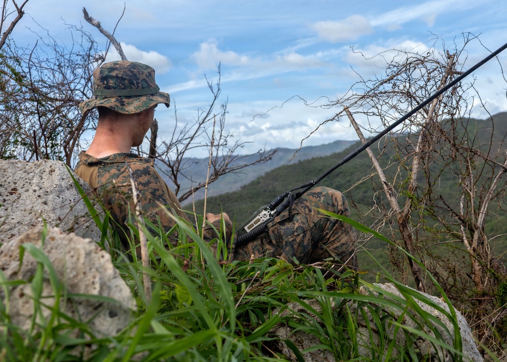 A military personnel in digital camouflage uniform and cover (hat) is positioned on a rocky elevated outcrop overlooking a mountainous, forested landscape. The individual is holding or operating a military radio with an extended antenna, suggesting a communications or observation role. The setting appears to be a field training exercise or operational deployment in a tropical or subtropical environment.