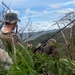 A military personnel in digital camouflage uniform and cover (hat) is positioned on a rocky elevated outcrop overlooking a mountainous, forested landscape. The individual is holding or operating a military radio with an extended antenna, suggesting a communications or observation role. The setting appears to be a field training exercise or operational deployment in a tropical or subtropical environment.