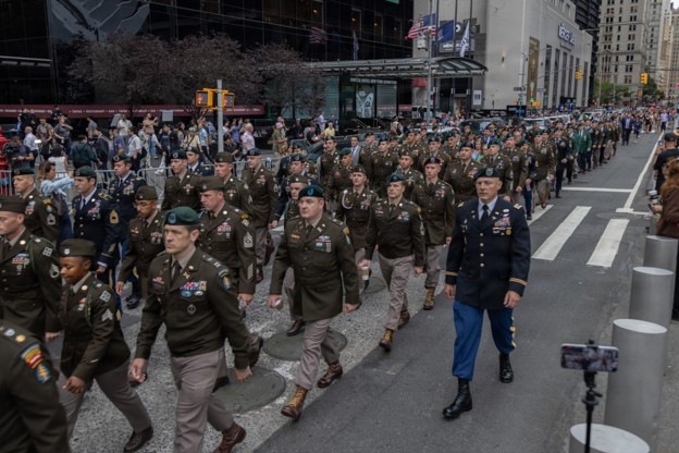 A large military parade is taking place on a city street, likely in New York City based on the urban architecture and building signage visible in the background. Numerous soldiers in U.S. Army dress uniforms, including olive drab service uniforms and green berets, are marching in formation down the street. A senior officer in a blue dress uniform is visible on the right side, and spectators line the sidewalks.