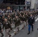 A large military parade is taking place on a city street, likely in New York City based on the urban architecture and building signage visible in the background. Numerous soldiers in U.S. Army dress uniforms, including olive drab service uniforms and green berets, are marching in formation down the street. A senior officer in a blue dress uniform is visible on the right side, and spectators line the sidewalks.