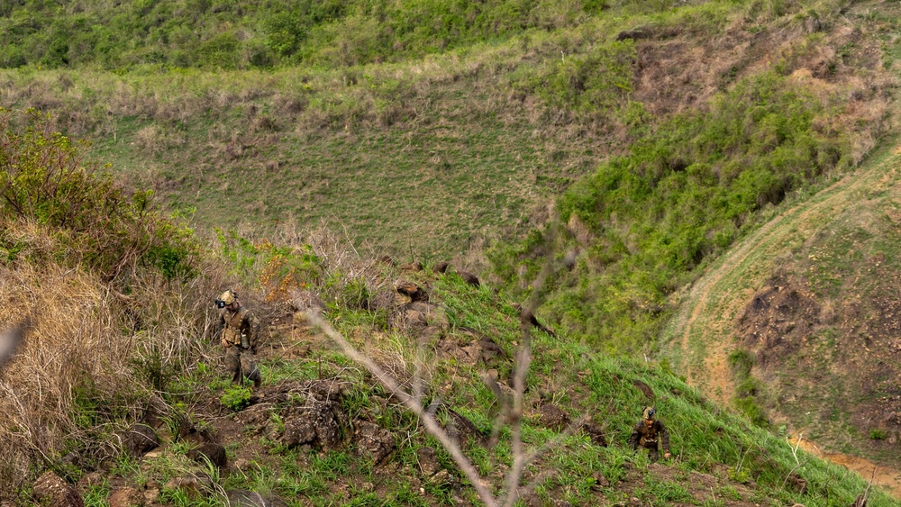 Two armed military personnel in camouflage gear are navigating a steep, rocky hillside terrain covered with grass and sparse vegetation. The soldiers appear to be conducting a patrol or training exercise on a rugged, hilly landscape. The image is taken from an elevated vantage point, looking down into a valley-like depression between hills.