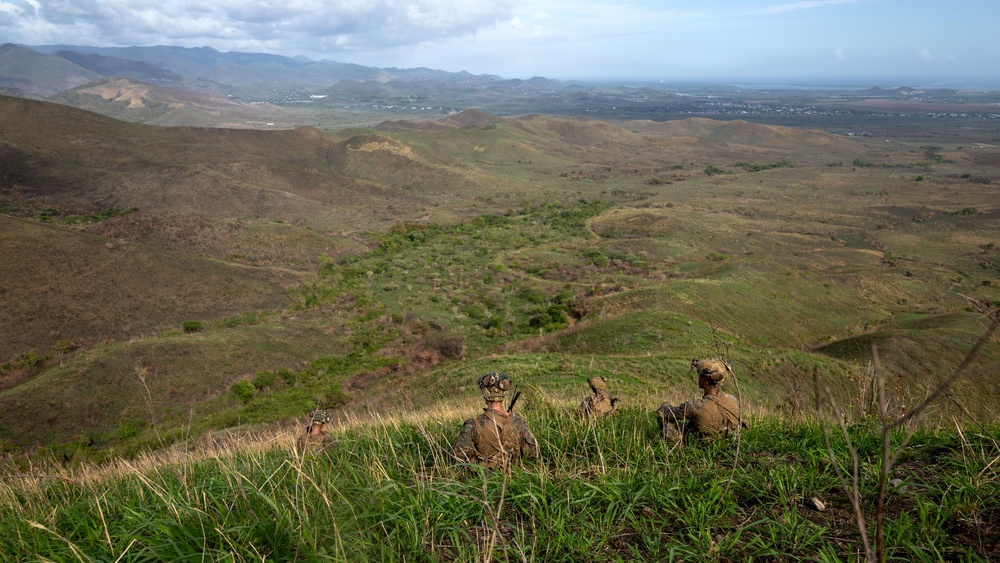 Three military personnel in combat gear are positioned on a hilltop overlooking a vast rolling landscape with mountains in the background. They appear to be in an observation or overwatch position, surveying the terrain below. The setting suggests a training exercise or tactical reconnaissance operation in a semi-arid, grassy highland environment.