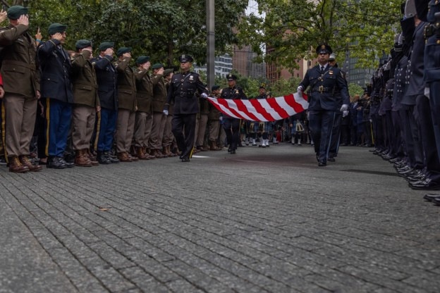 A formal ceremonial procession is taking place along a brick-paved pathway, with two officers in dark uniforms carrying a folded American flag between two lines of saluting personnel. The left side features individuals in olive/brown military-style uniforms with green berets, while the right side shows personnel in dark blue uniforms. The setting appears to be an outdoor memorial or ceremonial venue surrounded by trees and urban buildings.