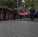 A formal ceremonial procession is taking place along a brick-paved pathway, with two officers in dark uniforms carrying a folded American flag between two lines of saluting personnel. The left side features individuals in olive/brown military-style uniforms with green berets, while the right side shows personnel in dark blue uniforms. The setting appears to be an outdoor memorial or ceremonial venue surrounded by trees and urban buildings.