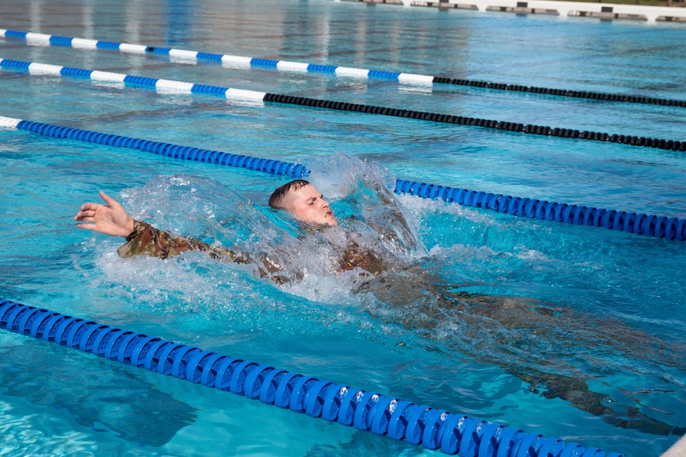 A person wearing camouflage military-style clothing is swimming backstroke in a competitive outdoor swimming pool. The swimmer is captured mid-stroke with significant water splash around them, suggesting vigorous movement. The pool features standard lane dividers in blue and white, with multiple lanes visible.
