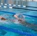 A person wearing camouflage military-style clothing is swimming backstroke in a competitive outdoor swimming pool. The swimmer is captured mid-stroke with significant water splash around them, suggesting vigorous movement. The pool features standard lane dividers in blue and white, with multiple lanes visible.