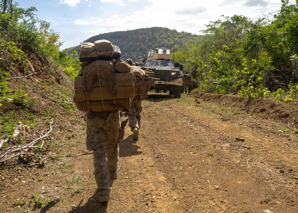 A column of military personnel in full combat gear and large rucksacks advances on foot along a dirt trail in a tropical or subtropical environment. An armored military vehicle, likely an MRAP or similar wheeled armored vehicle in tan/desert coloring, follows behind the dismounted troops. The scene appears to depict a patrol or tactical movement exercise in a hilly, vegetated terrain.