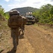 A column of military personnel in full combat gear and large rucksacks advances on foot along a dirt trail in a tropical or subtropical environment. An armored military vehicle, likely an MRAP or similar wheeled armored vehicle in tan/desert coloring, follows behind the dismounted troops. The scene appears to depict a patrol or tactical movement exercise in a hilly, vegetated terrain.