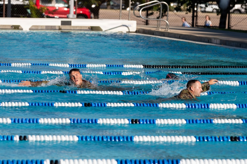 Two military personnel in full camouflage uniforms are swimming in adjacent lanes of an outdoor competition pool. The swimmers appear to be participating in a military fitness or assessment exercise, as evidenced by their combat uniforms worn in the water. Spectators can be seen seated in the background beyond the pool fence.
