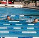 Two military personnel in full camouflage uniforms are swimming in adjacent lanes of an outdoor competition pool. The swimmers appear to be participating in a military fitness or assessment exercise, as evidenced by their combat uniforms worn in the water. Spectators can be seen seated in the background beyond the pool fence.
