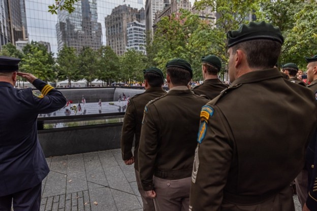 A solemn memorial ceremony is taking place at the 9/11 Memorial reflecting pool in New York City. Military personnel in olive/dark green uniforms with green berets and a uniformed officer in dark blue rendering a salute are gathered at the edge of the memorial pool. Small American flags and flowers are visible placed along the memorial's bronze parapet inscribed with victims' names.