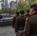 A solemn memorial ceremony is taking place at the 9/11 Memorial reflecting pool in New York City. Military personnel in olive/dark green uniforms with green berets and a uniformed officer in dark blue rendering a salute are gathered at the edge of the memorial pool. Small American flags and flowers are visible placed along the memorial's bronze parapet inscribed with victims' names.