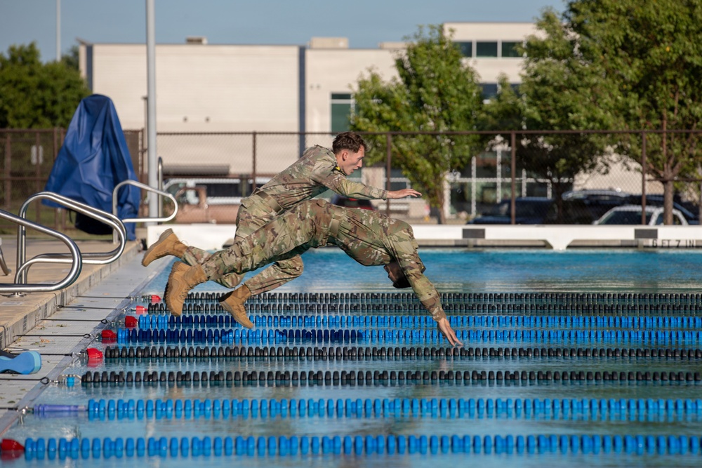 Two military personnel in full camouflage uniforms and combat boots are captured mid-air as they jump or dive into an outdoor swimming pool. They appear to be conducting water survival or combat swimming training, fully clothed in their operational camouflage pattern (OCP) uniforms. The setting is a regulation outdoor lap pool at what appears to be a military installation.