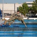 Two military personnel in full camouflage uniforms and combat boots are captured mid-air as they jump or dive into an outdoor swimming pool. They appear to be conducting water survival or combat swimming training, fully clothed in their operational camouflage pattern (OCP) uniforms. The setting is a regulation outdoor lap pool at what appears to be a military installation.
