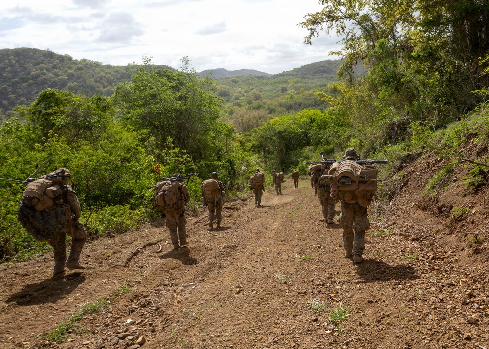 A column of military personnel in full combat gear and camouflage uniforms patrol along a dirt trail through a tropical or subtropical hilly landscape. The soldiers are carrying large rucksacks and visible weapons, moving in a single-file or staggered formation away from the camera. The terrain features red-brown soil, dense green vegetation, and rolling hills in the background.