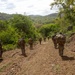 A column of military personnel in full combat gear and camouflage uniforms patrol along a dirt trail through a tropical or subtropical hilly landscape. The soldiers are carrying large rucksacks and visible weapons, moving in a single-file or staggered formation away from the camera. The terrain features red-brown soil, dense green vegetation, and rolling hills in the background.