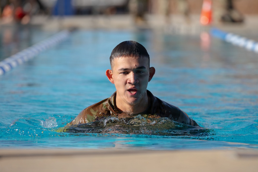 A young male in military camouflage uniform is swimming in an outdoor pool, with his head and upper torso visible above the water. His hair and uniform are wet, and his mouth is slightly open, suggesting physical exertion. This appears to be a military water training or swim qualification exercise.