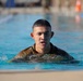 A young male in military camouflage uniform is swimming in an outdoor pool, with his head and upper torso visible above the water. His hair and uniform are wet, and his mouth is slightly open, suggesting physical exertion. This appears to be a military water training or swim qualification exercise.