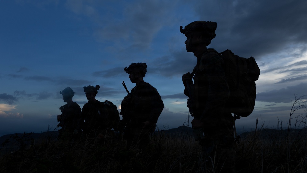 Four heavily equipped military personnel are silhouetted against a twilight sky, standing in a line on elevated terrain with sparse dry grass. The image is taken during dusk or dawn, creating a dramatic dark silhouette effect against blue-toned clouds. The figures appear to be on patrol or at a tactical observation point.