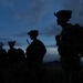 Four heavily equipped military personnel are silhouetted against a twilight sky, standing in a line on elevated terrain with sparse dry grass. The image is taken during dusk or dawn, creating a dramatic dark silhouette effect against blue-toned clouds. The figures appear to be on patrol or at a tactical observation point.