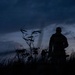 A silhouetted figure stands in an outdoor field or grassland during twilight or dusk, facing away from the camera. The dramatic sky features heavy, dark storm clouds with faint pink and blue hues near the horizon. Wild grasses and plants are visible in the foreground, creating a moody, atmospheric composition.