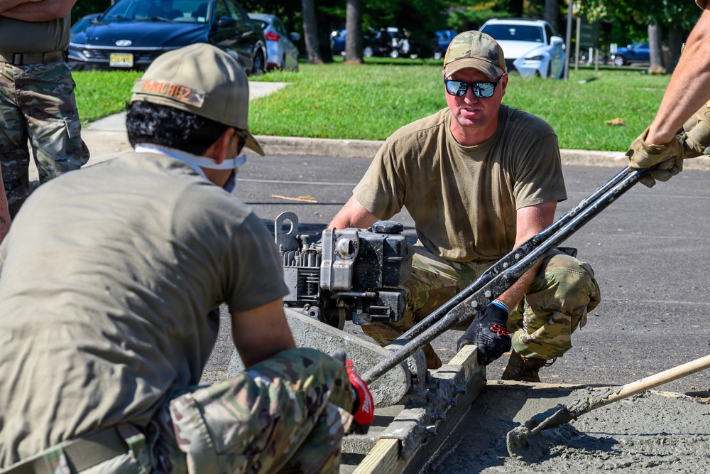 Military personnel in camouflage uniforms are working on a concrete construction or repair project in what appears to be a parking lot area. At least three service members are visible, using a concrete screed or vibrator machine to level fresh concrete within wooden form boards. The setting appears to be a military installation or government facility based on the uniforms and surrounding infrastructure.