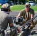Military personnel in camouflage uniforms are working on a concrete construction or repair project in what appears to be a parking lot area. At least three service members are visible, using a concrete screed or vibrator machine to level fresh concrete within wooden form boards. The setting appears to be a military installation or government facility based on the uniforms and surrounding infrastructure.