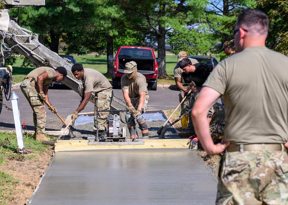 A group of military personnel in camouflage uniforms are pouring and finishing a concrete sidewalk or pathway in what appears to be a base or installation setting. Several soldiers are actively working with shovels and a concrete screed/vibrator machine to level and smooth the freshly poured concrete. A supervisor or observer in the foreground watches the work progress from behind.