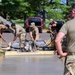 A group of military personnel in camouflage uniforms are pouring and finishing a concrete sidewalk or pathway in what appears to be a base or installation setting. Several soldiers are actively working with shovels and a concrete screed/vibrator machine to level and smooth the freshly poured concrete. A supervisor or observer in the foreground watches the work progress from behind.