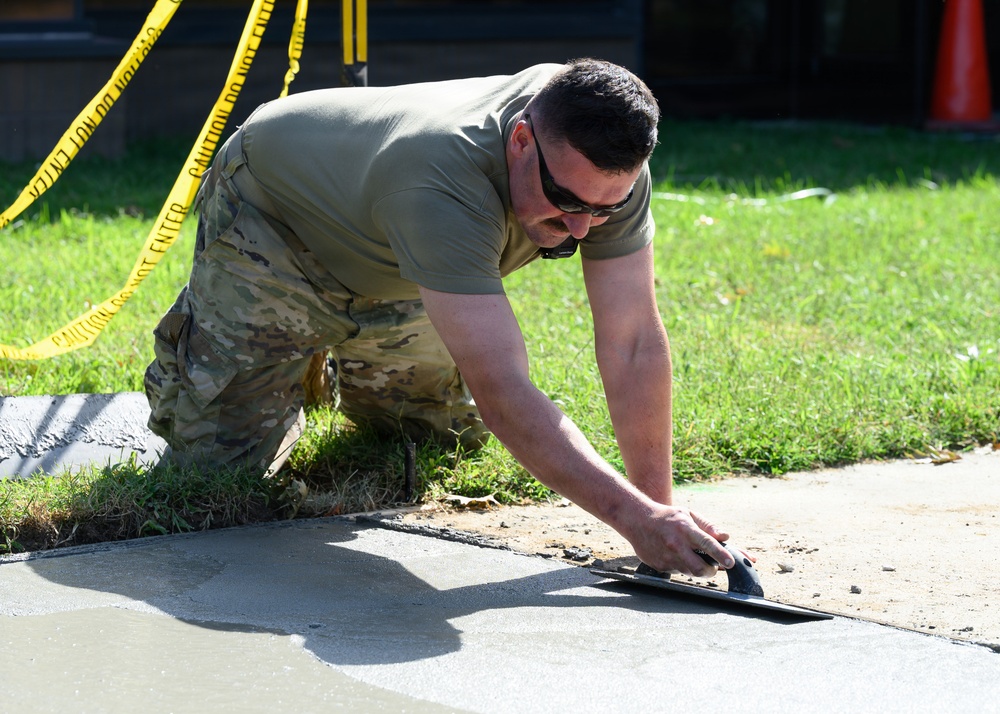 A person wearing military-style camouflage pants and an olive drab t-shirt is kneeling on grass while smoothing freshly poured concrete with a hand float/trowel. The work area is cordoned off with yellow caution tape visible in the background. The individual is wearing sunglasses and appears to be performing concrete finishing work on what looks like a sidewalk or pathway.