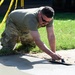 A person wearing military-style camouflage pants and an olive drab t-shirt is kneeling on grass while smoothing freshly poured concrete with a hand float/trowel. The work area is cordoned off with yellow caution tape visible in the background. The individual is wearing sunglasses and appears to be performing concrete finishing work on what looks like a sidewalk or pathway.