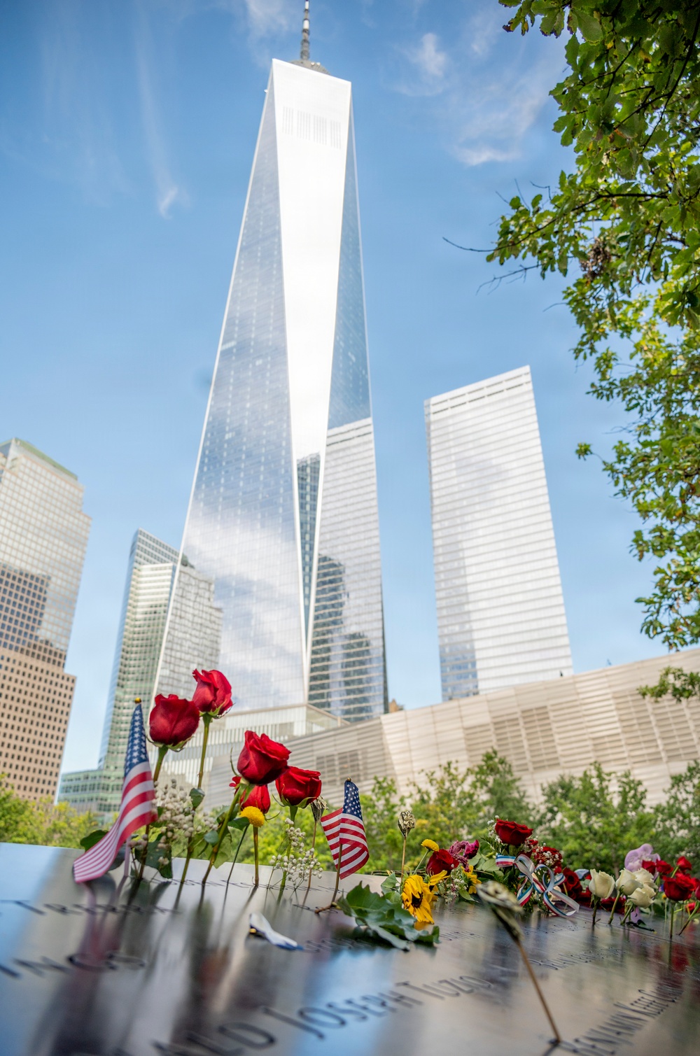 The image shows the 9/11 Memorial in Lower Manhattan, New York City, with One World Trade Center (Freedom Tower) prominently visible in the background. The foreground features the bronze memorial parapet with engraved names, adorned with red roses, mixed flowers, and small American flags placed as tributes. The scene conveys solemn remembrance against a clear blue sky with surrounding skyscrapers.