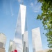 The image shows the 9/11 Memorial in Lower Manhattan, New York City, with One World Trade Center (Freedom Tower) prominently visible in the background. The foreground features the bronze memorial parapet with engraved names, adorned with red roses, mixed flowers, and small American flags placed as tributes. The scene conveys solemn remembrance against a clear blue sky with surrounding skyscrapers.